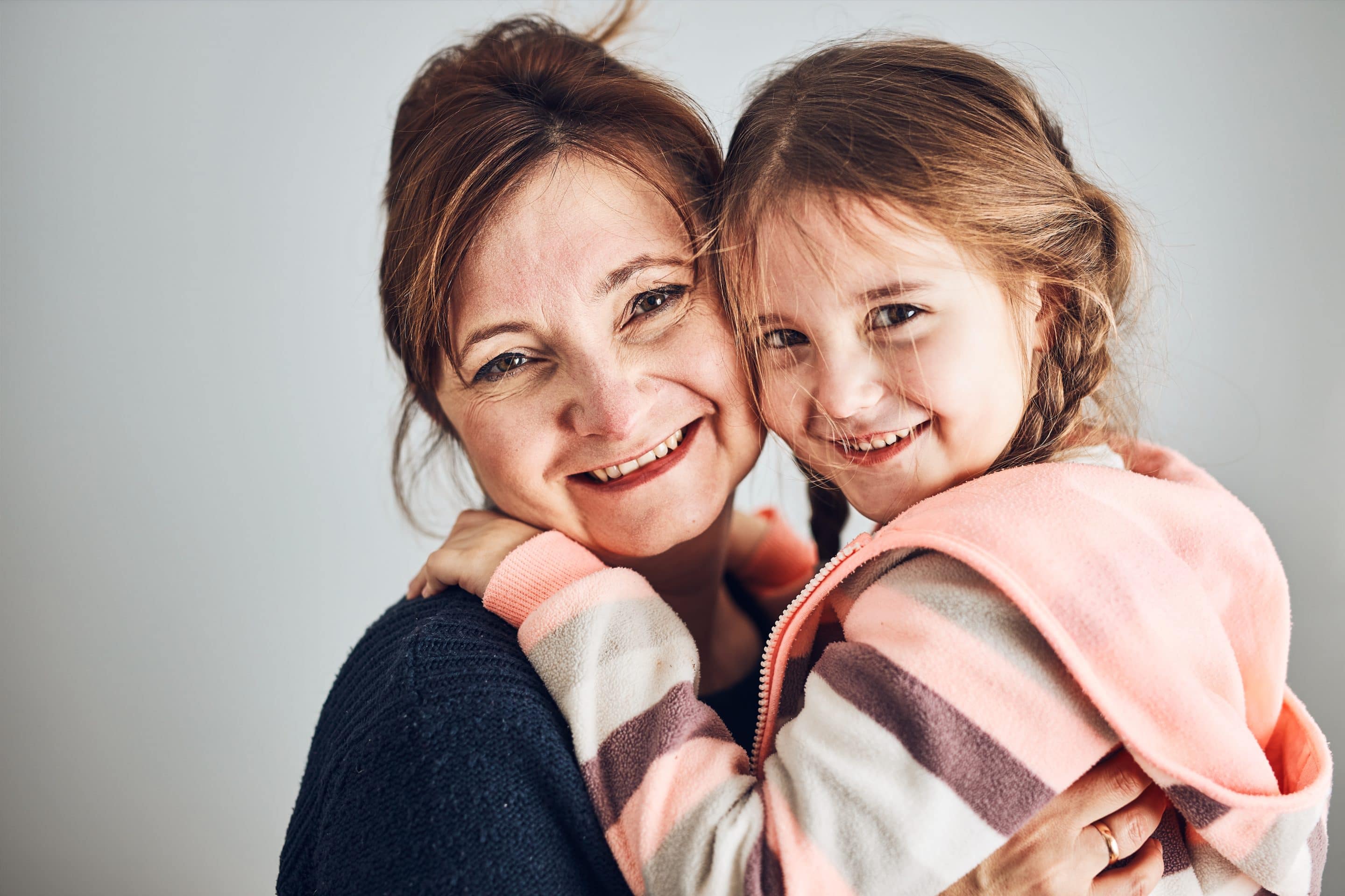 Happy mother and daughter embracing, hugging and smiling together. Family portrait. Happy moment. Woman and little girl posing to camera. Grey background